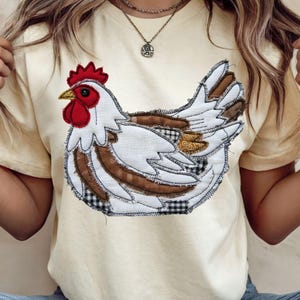 May include: Cream-colored t-shirt featuring a detailed appliqué of a chicken. The chicken has white feathers, brown and black accents, a red comb and wattle, and a yellow beak. The shirt is a casual style.