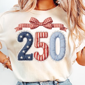 May include: Cream-coloured t-shirt with the number "250" in a patriotic design. The "2" is blue with white stars, the "5" is red and white striped, and the "0" is blue gingham. A red and white plaid bow and ribbon are above the numbers.