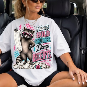 K&ouml;nnte beinhalten: Ein wei&szlig;es T-Shirt mit einer Grafik eines Waschb&auml;ren, der eine rosa Sonnenbrille tr&auml;gt und einen langen Kassenbon h&auml;lt. Der Text lautet, I don't hold grudges. I keep receipts. Das Design enth&auml;lt rosa Blitze, Karomuster und verstreutes Papier.