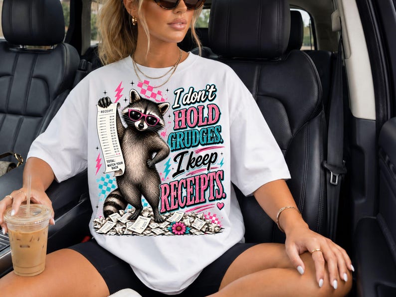 K&ouml;nnte beinhalten: Ein wei&szlig;es T-Shirt mit einer Grafik eines Waschb&auml;ren, der eine rosa Sonnenbrille tr&auml;gt und einen langen Kassenbon h&auml;lt. Der Text lautet, I don't hold grudges. I keep receipts. Das Design enth&auml;lt rosa Blitze, Karomuster und verstreutes Papier.