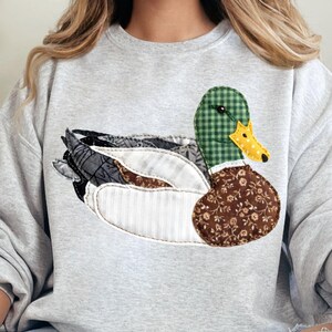 May include: A light gray sweatshirt featuring a patchwork duck design. The duck has a green checkered head, a yellow dotted beak, and a brown floral patterned body. The wings are a mix of patterned fabrics.