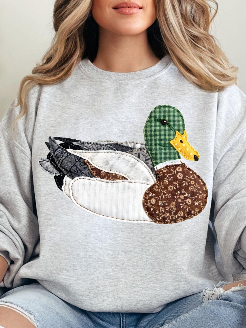 May include: A light gray sweatshirt featuring a patchwork duck design. The duck has a green checkered head, a yellow dotted beak, and a brown floral patterned body. The wings are a mix of patterned fabrics.
