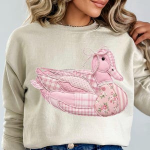 May include: Beige sweatshirt featuring a large, appliqued pink duck design. The duck is composed of various pink patterned fabrics, including plaid, polka dots, and floral prints. A bow adorns the duck's head.