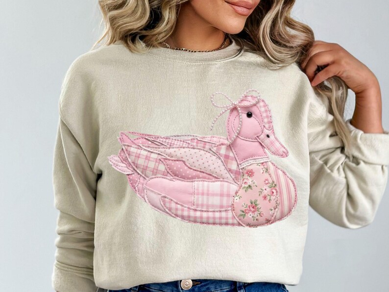 May include: Beige sweatshirt featuring a large, appliqued pink duck design. The duck is composed of various pink patterned fabrics, including plaid, polka dots, and floral prints. A bow adorns the duck's head.
