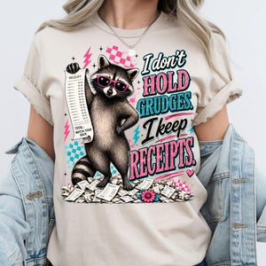 K&ouml;nnte beinhalten: Ein beiges T-Shirt mit einer Grafik eines Waschb&auml;ren, der eine rosa Sonnenbrille tr&auml;gt, einen langen Kassenbon h&auml;lt und auf einem Haufen Kassenbons steht. Der Text lautet, I don't HOLD GRUDGES. I keep RECEIPTS. Das Design enth&auml;lt rosa Blitze und Karomuster.