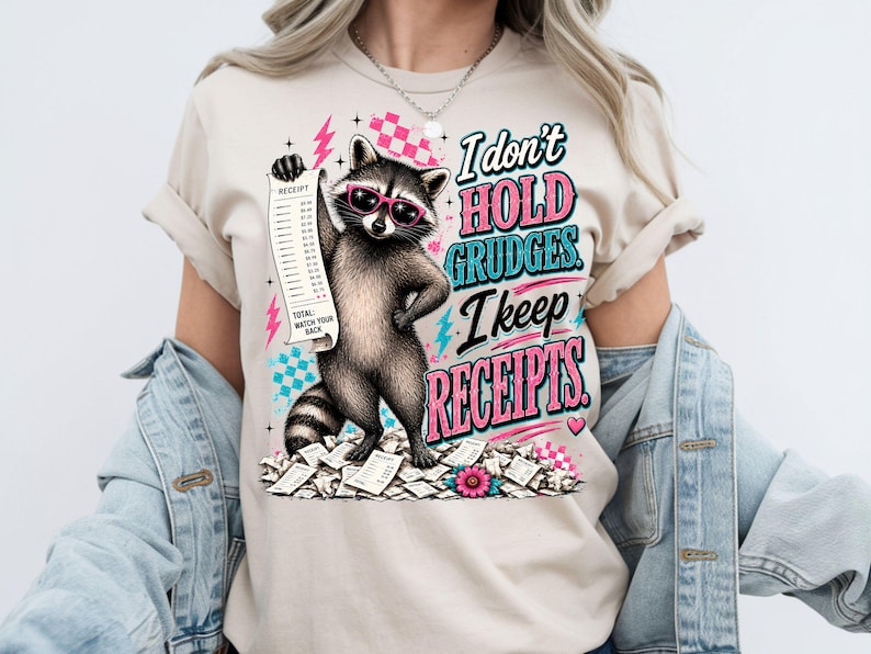 K&ouml;nnte beinhalten: Ein beiges T-Shirt mit einer Grafik eines Waschb&auml;ren, der eine rosa Sonnenbrille tr&auml;gt, einen langen Kassenbon h&auml;lt und auf einem Haufen Kassenbons steht. Der Text lautet, I don't HOLD GRUDGES. I keep RECEIPTS. Das Design enth&auml;lt rosa Blitze und Karomuster.