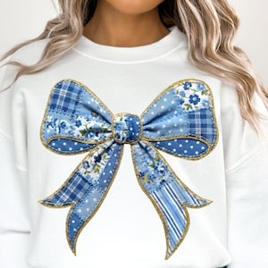 Könnte beinhalten: Weißes Sweatshirt mit einem großen Patchwork-Schleifendesign. Der Bogen enthält blaue Blumen-, Tupfen- und Karomuster mit goldenen Verzierungen. Ein einzigartiges und stilvolles Kleidungsstück.