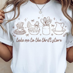May include: White t-shirt with a brown line drawing of kitchen items, including stacked teacups, a hen in a bowl, utensils in a jar, and a pitcher. The text "take me to the thrift store" is printed below the illustrations.