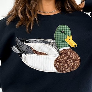 May include: Black sweatshirt featuring a patchwork duck design. The duck has a green checkered head, a yellow and white polka-dotted beak, and a brown floral patterned body. The wings are made of various patterned fabrics.