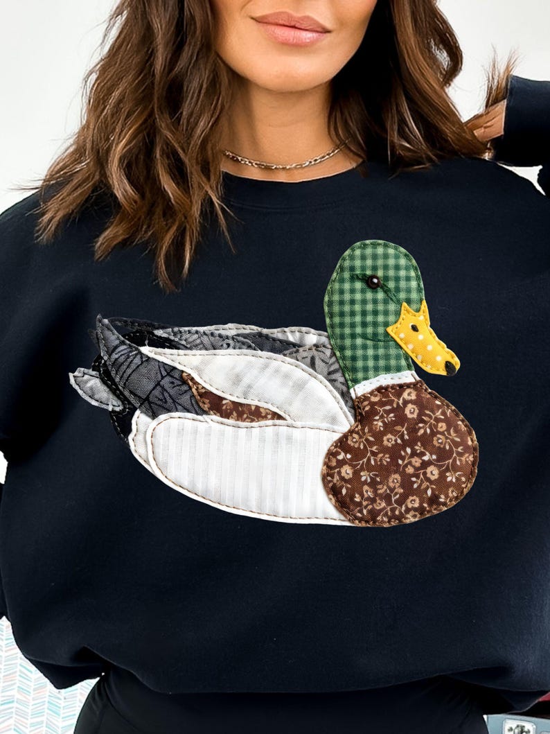May include: Black sweatshirt featuring a patchwork duck design. The duck has a green checkered head, a yellow and white polka-dotted beak, and a brown floral patterned body. The wings are made of various patterned fabrics.