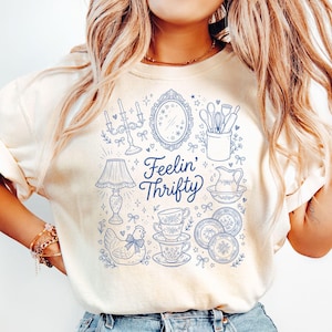 Op de afbeelding: Crèmekleurig T-shirt met een blauwe illustratie van vintage keuken- en woondecoratie-items. Het ontwerp bevat een lamp, spiegel, kopjes en de tekst "Feelin' Thrifty".
