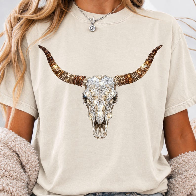 May include: A beige short-sleeve t-shirt featuring a large graphic of a bull skull. The skull is decorated with clear, sparkling rhinestones, while the long, curved horns are adorned with a mix of brown, amber, and gold-toned crystals.