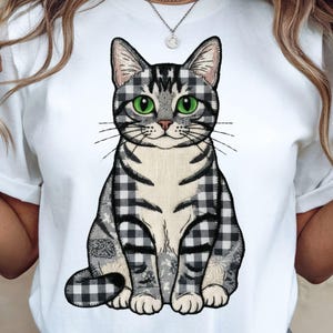 Puede incluir: Camiseta blanca con una ilustración de un gato sentado. El gato tiene un patrón de cuadros grises y negros con ojos verdes. Las patas y la cola del gato son visibles, y el diseño está centrado en la camiseta.