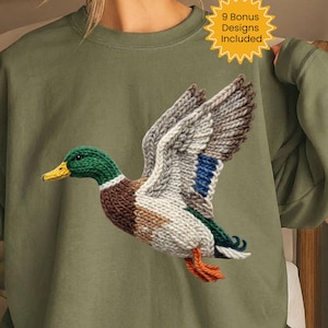 May include: Olive green sweatshirt featuring a detailed knitted duck design in flight. The duck has a green head, brown body, and white and blue wings. The text "9 Bonus Designs Included" is in a yellow starburst.