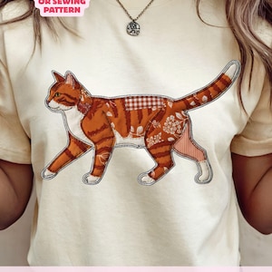 Op de afbeelding: Crèmekleurig T-shirt met een kattenontwerp in patchwork-stijl in tinten oranje, wit en rood. De kat heeft een gedetailleerd patroon en is omlijnd met een zwarte steek. De tekst op het shirt luidt "NOT A CUTTING OR SEWING PATTERN" en "DIGITAL DOWNLOAD - NOT A PHYSICAL ITEM."