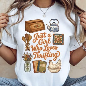 Puede incluir: Camiseta blanca con el texto "Just a Girl Who Loves Thrifting" en escritura naranja. La camiseta presenta ilustraciones de una panera, tetera, cucharas de madera, un edredón, flores, libros de cocina y una gallina.