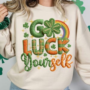 Puede incluir: Sudadera color crema con la frase bordada "Go Luck Yourself" en verde y naranja. El diseño incluye un trébol, un arcoíris y estrellas, ideal para el Día de San Patricio.