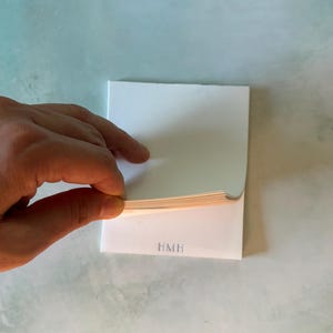 May include: A rectangular white notepad with a stack of paper sheets. The bottom sheet features the letters HMH printed in a simple gray font. The top sheet is being lifted to reveal the thickness of the paper stack underneath.