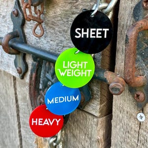 Pode incluir: Quatro etiquetas redondas, codificadas por cores, presas a uma corrente. As etiquetas são pretas, verdes, azuis e vermelhas. A etiqueta preta diz "SHEET", a etiqueta verde diz "LIGHT WEIGHT", a etiqueta azul diz "MEDIUM" e a etiqueta vermelha diz "HEAVY".