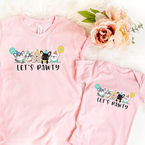 Pode incluir: Tr&ecirc;s camisetas rosa e um body com um design de festa canina. As camisetas e o body t&ecirc;m o texto "Let's Pawty" e apresentam c&atilde;es de desenho animado com chap&eacute;us de festa e bal&otilde;es. As camisetas est&atilde;o em v&aacute;rios tamanhos.