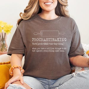 May include: A brown t-shirt with the word "PROCRASTIBAKING" and a rolling pin graphic. The definition of the word is printed below the graphic. The person is wearing ripped blue jeans and holding a white mug with a floral design.