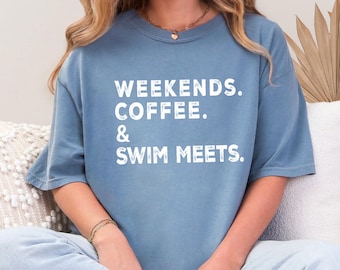 Peut inclure: Un t-shirt bleu avec les mots "WEEKENDS. COFFEE. & SWIM MEETS." en lettres blanches. Le t-shirt est de coupe décontractée et est porté par une personne assise sur un canapé.