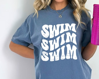 Peut inclure: Un t-shirt bleu jean avec les mots "Swim Swim Swim" en blanc, dans une police de caractères rétro. Le t-shirt est porté par une personne tenant un gobelet rose.