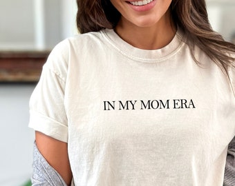 Może przedstawiać: Biały t-shirt z nadrukiem "IN MY MOM ERA" w kolorze czarnym na przodzie. Koszulkę nosi osoba w niebieskich dżinsach i szarym kardiganie.
