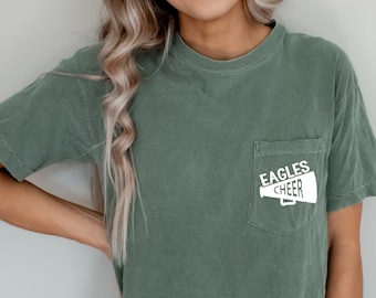 Pode incluir: Uma camiseta verde musgo com um bolso branco que mostra um megafone com o texto "Eagles Cheer".