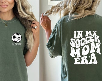 Könnte beinhalten: Moosgrünes T-Shirt mit einem Fußball-Grafik und dem Namen "Jackson" darunter. Auf der Rückseite des Shirts steht "IN MY SOCCER MOM ERA" in weißen Retro-Buchstaben. Ein lässiges, bequemes T-Shirt.