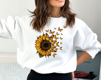 Könnte beinhalten: Weißer Pullover mit einem Sonnenblumen- und Schmetterlingsdruck. Die Sonnenblume ist gelb mit brauner Mitte und grünen Blättern. Die Schmetterlinge sind gelb.