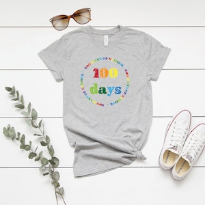Puede incluir: Una camiseta gris jaspeado de manga corta con un estampado gr&aacute;fico colorido. El dise&ntilde;o incluye el texto 100 days en letras grandes y multicolores, rodeado por un borde circular con el texto repetido 100 DAYS OF SCHOOL en colores arco&iacute;ris.