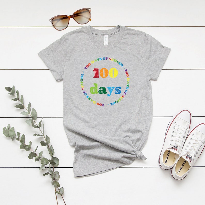 Puede incluir: Una camiseta gris jaspeado de manga corta con un estampado gr&aacute;fico colorido. El dise&ntilde;o incluye el texto 100 days en letras grandes y multicolores, rodeado por un borde circular con el texto repetido 100 DAYS OF SCHOOL en colores arco&iacute;ris.