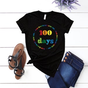 Puede incluir: Una camiseta negra de manga corta con un estampado gr&aacute;fico colorido. El dise&ntilde;o incluye el texto 100 days en letras grandes y multicolores, rodeado por un borde circular con el texto repetido 100 DAYS OF SCHOOL en colores del arco&iacute;ris.