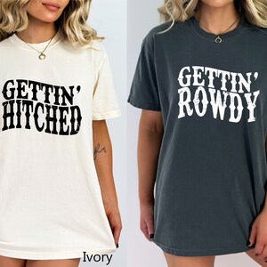 May include: Two women wearing oversized t-shirts. The shirt on the left is ivory with black text that says "Gettin' Hitched". The shirt on the right is dark gray with white text that says "Gettin' Rowdy".
