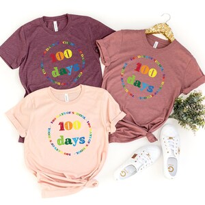 Puede incluir: Tres camisetas de manga corta y cuello redondo en color granate, malva y melocot&oacute;n claro. Cada camiseta presenta un gr&aacute;fico circular con el texto 100 DAYS OF SCHOOL en letras de colores del arco&iacute;ris, con el n&uacute;mero 100 y la palabra days centrados.