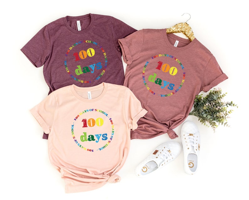 Puede incluir: Tres camisetas de manga corta y cuello redondo en color granate, malva y melocot&oacute;n claro. Cada camiseta presenta un gr&aacute;fico circular con el texto 100 DAYS OF SCHOOL en letras de colores del arco&iacute;ris, con el n&uacute;mero 100 y la palabra days centrados.