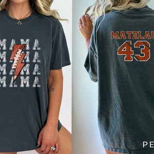 May include: A dark gray t-shirt with a white lightning bolt graphic and the word "MAMA" repeated five times. The back of the shirt has the name "MATELAU" and the number "43" in orange and black.