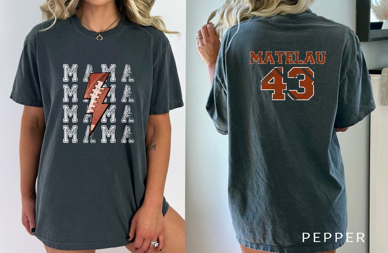 May include: A dark gray t-shirt with a white lightning bolt graphic and the word "MAMA" repeated five times. The back of the shirt has the name "MATELAU" and the number "43" in orange and black.