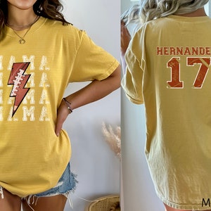 May include: A mustard yellow t-shirt with a football and lightning bolt graphic on the front and the text "MAMA" repeated four times. The back of the shirt has the text "HERNANDEZ" and the number "17" in a distressed style.