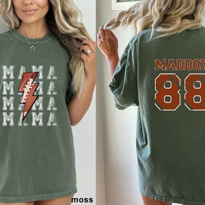 May include: A green t-shirt with a white lightning bolt design featuring a football. The back of the shirt has the name Maddox and the number 88 in orange.