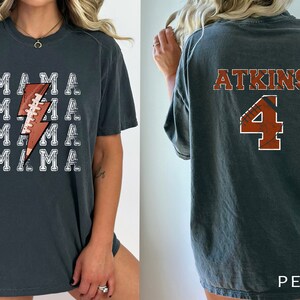 May include: A dark gray t-shirt with a white lightning bolt design featuring a football. The text "MAMA" is repeated four times on the shirt. The back of the shirt has the text "ATKINS" and the number "4" in a football design.