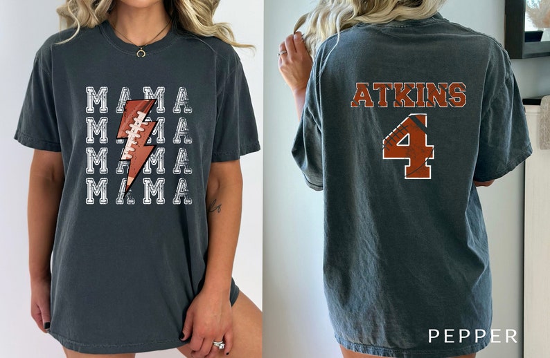 May include: A dark gray t-shirt with a white lightning bolt design featuring a football. The text "MAMA" is repeated four times on the shirt. The back of the shirt has the text "ATKINS" and the number "4" in a football design.