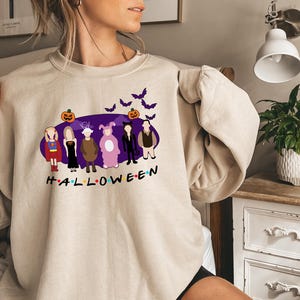 May include: A beige sweatshirt featuring a purple graphic of characters dressed in Halloween costumes. The graphic includes a pumpkin, bats, and the word "HALLOWEEN".