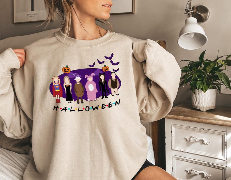 May include: A beige sweatshirt featuring a purple graphic of characters dressed in Halloween costumes. The graphic includes a pumpkin, bats, and the word "HALLOWEEN".