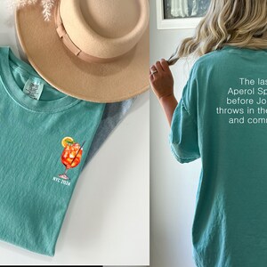May include: A teal blue t-shirt with a white graphic of a cocktail drink and the text "NYC 2023". The back of the shirt has white text that reads "The last Aperol Spritz before Jordan throws in the towel and commits."