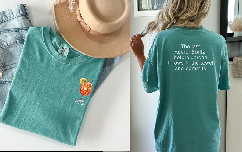 May include: A teal blue t-shirt with a white graphic of a cocktail drink and the text "NYC 2023". The back of the shirt has white text that reads "The last Aperol Spritz before Jordan throws in the towel and commits."