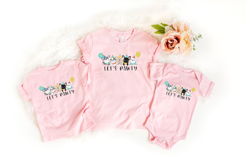 Pode incluir: Tr&ecirc;s camisetas rosa e um body com um design de festa canina. As camisetas e o body t&ecirc;m o texto "Let's Pawty" e apresentam c&atilde;es de desenho animado com chap&eacute;us de festa e bal&otilde;es. As camisetas est&atilde;o em v&aacute;rios tamanhos.