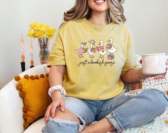 Puede incluir: Camiseta amarilla mostaza con un gráfico de cuatro gansos con gafas y libros. El texto "just a bookish goose" está debajo. La camiseta se usa con vaqueros azules.