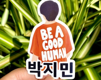 Puede incluir: Una pegatina con un personaje de dibujos animados que lleva una camisa naranja con el texto "Be a Good Human" en blanco. El texto "박지민" está escrito en negro debajo de la persona.